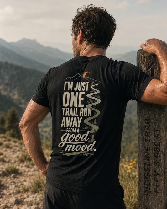 A man seen from behind wearing a black graphic t-shirt with a trail design and quote, leaning on a wooden Ridgeline Trail marker in a mountain landscape.