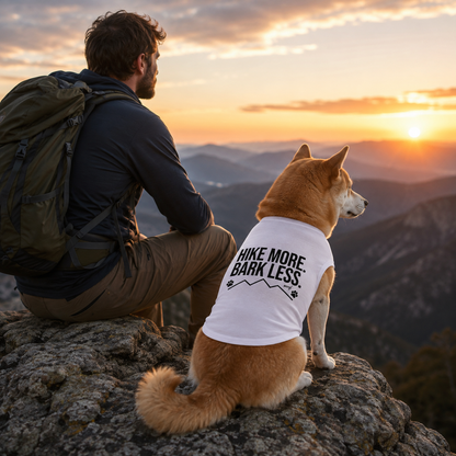 Shiba Inu dog wearing a white "Hike More Bark Less" shirt on a mountain at sunset.