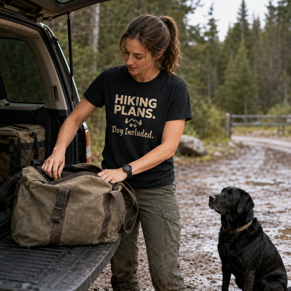 Woman in "Hiking Plans. Dog Included." shirt packs bag next to a black dog.