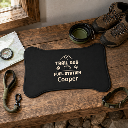 A black dog mat with white text reading 'TRAIL DOG FUEL STATION Cooper' and small mountain/paw/bowl graphics, styled on a rustic wooden bench with hiking shoes, a leash, collar, map, and cup nearby.