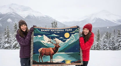 Two people holding a Banff National Park blanket in a snowy landscape with mountains.