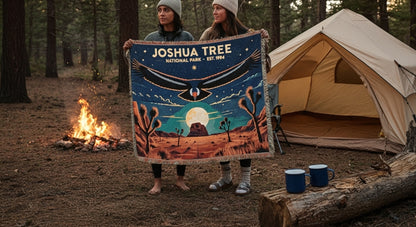Two people holding a 'Joshua Tree National Park' blanket near a campfire and tent.